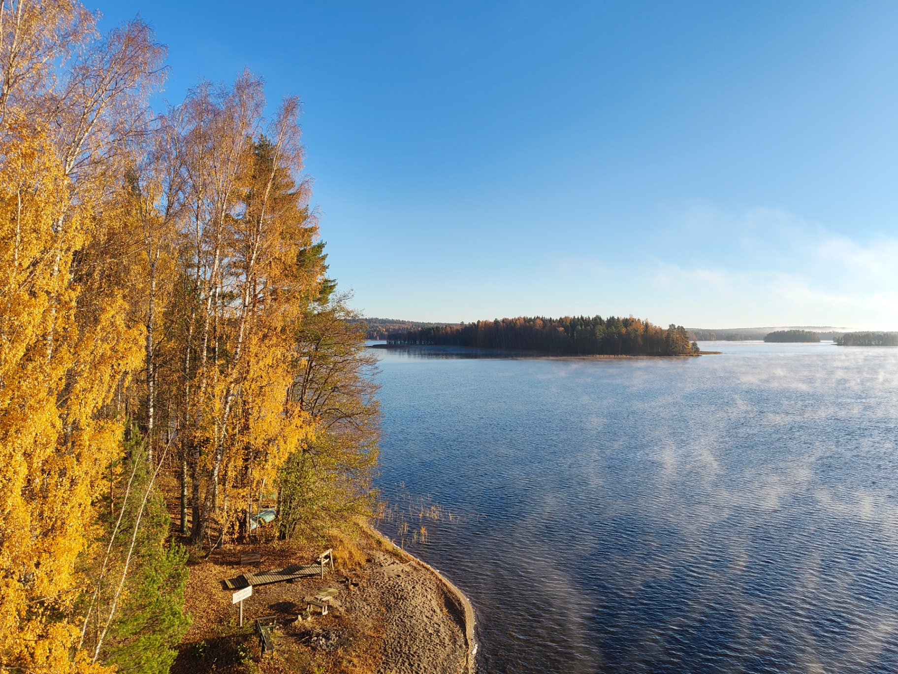 Sandy shore of Lake Päijänne below yellow birches, with a small bench, a wooden walkway and mist rising around a wooded island