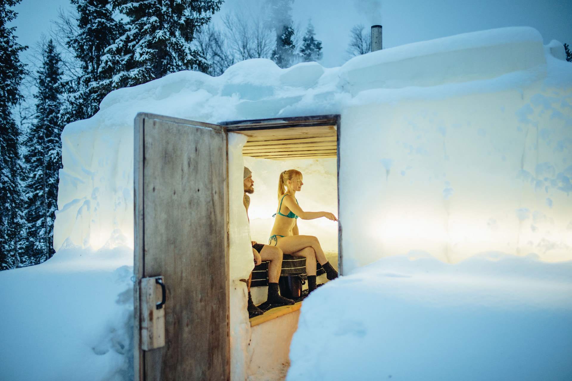 Two people sitting inside a warmly lit snow sauna with its wooden door open to a snowy Finnish forest