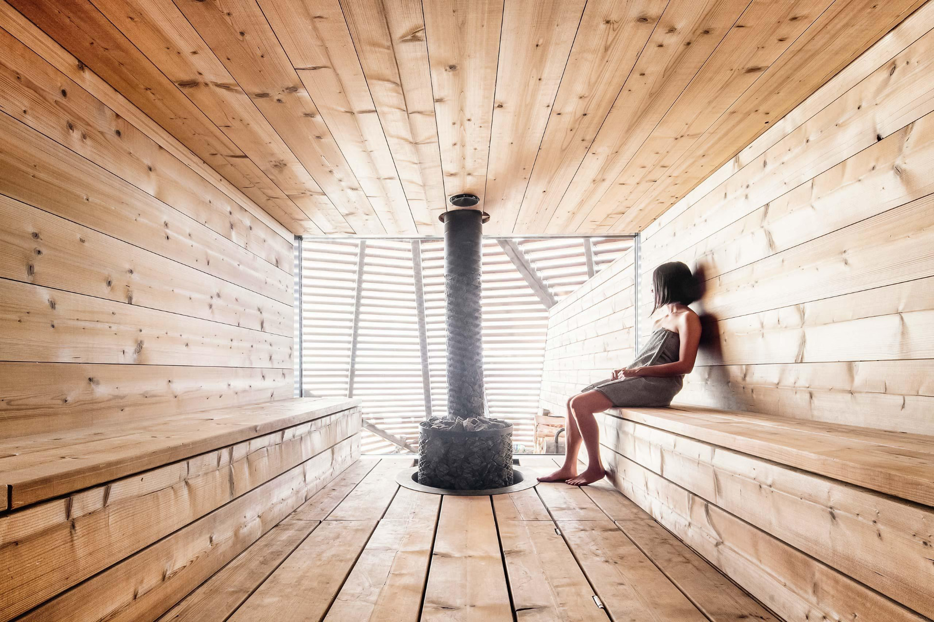A woman wrapped in a towel sitting on wooden sauna benches beside a stone-filled heater