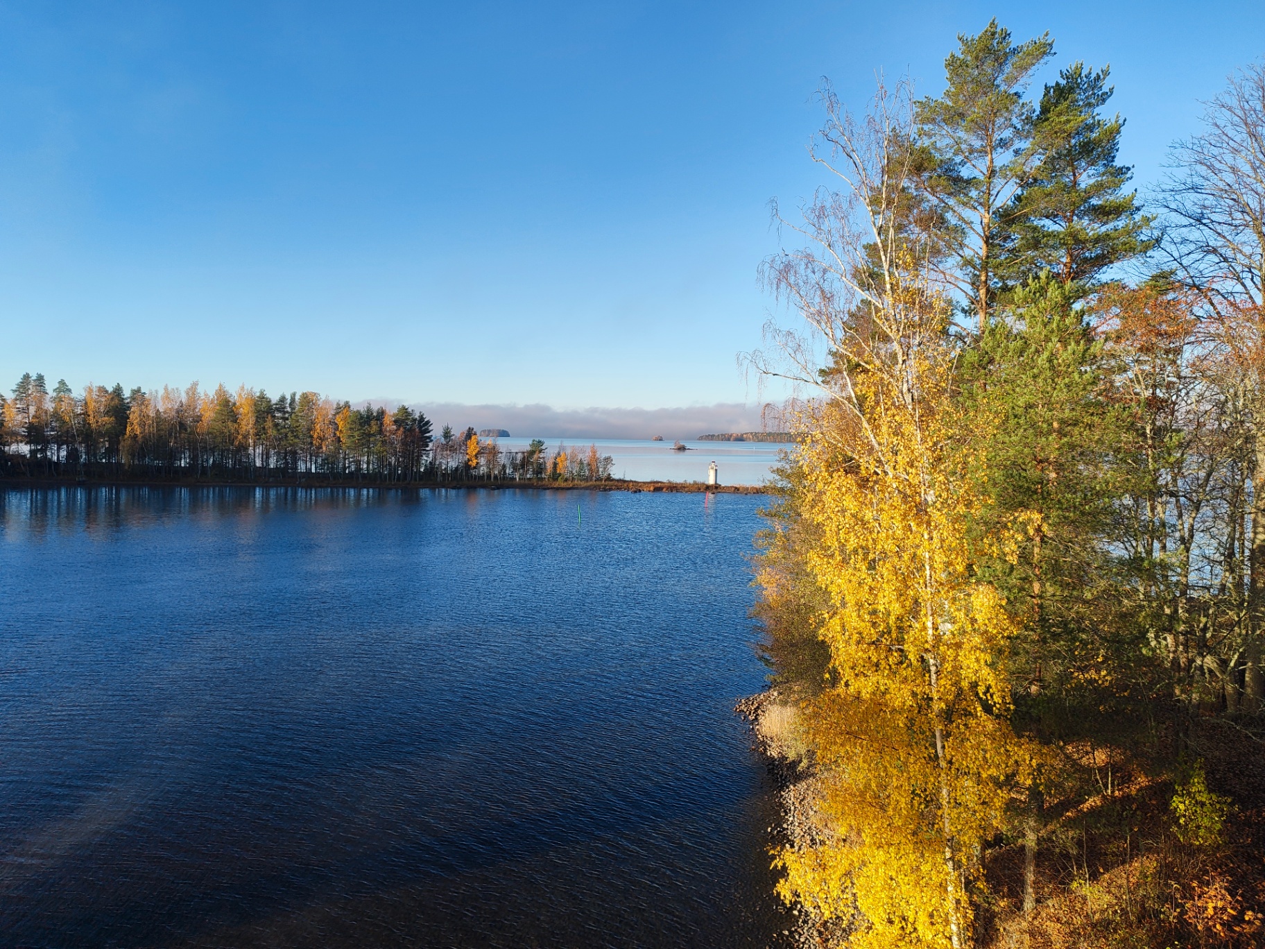 Raised view over Lake Päijänne in October: a wooded spit with yellow birches, a small white navigation light and mist on the open water